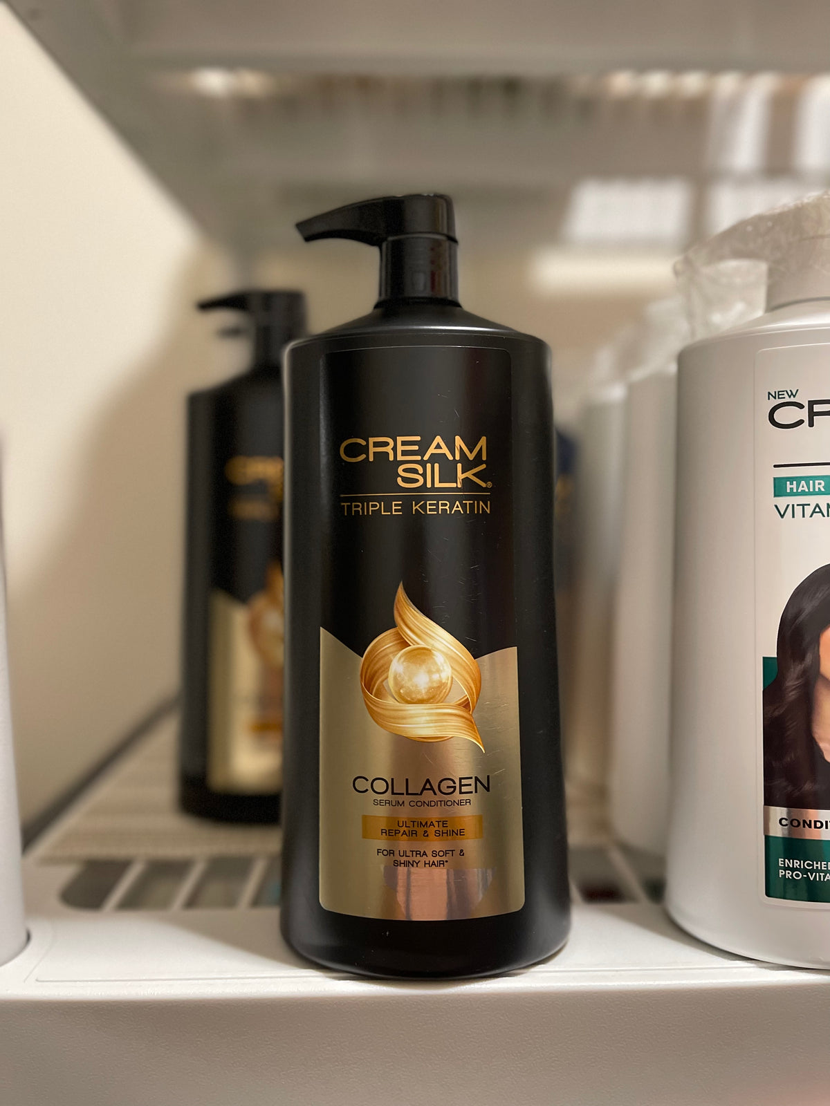 A bottle of Cream Silk Triple Keratin Rescue Conditioner with a pump dispenser, positioned on a shelf with other hair care products in the background.
